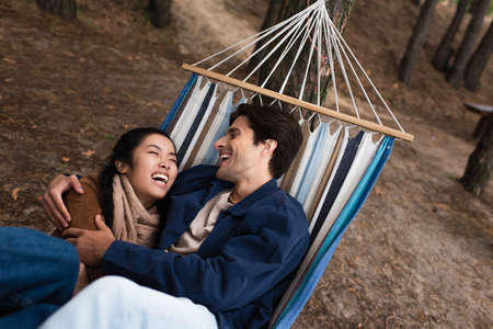 Excited multiethnic couple hugging in hammock during weekend