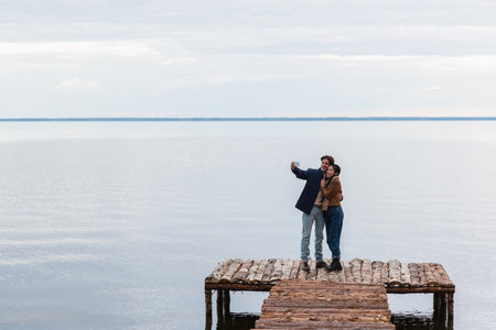 Happy multiethnic couple taking selfie on cellphone on pier near sea