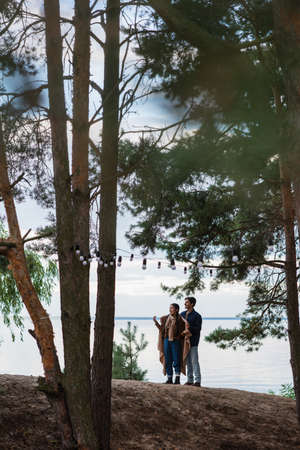 Smiling asian woman holding blanket near boyfriend on hillの写真素材