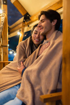 Smiling asian woman in blanket hugging boyfriend on glamping terraceの写真素材