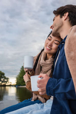 Man holding cup near smiling asian woman and lake during weekendの写真素材