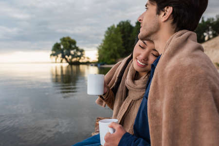 Smiling asian woman with closed eyes holding cup near boyfriend and lakeの写真素材