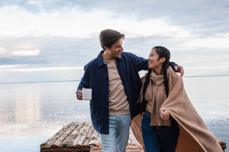 Smiling man with cup hugging asian girlfriend in blanket on pierの写真素材