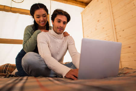 Young asian woman hugging boyfriend with blurred laptop on bed in glamping houseの写真素材