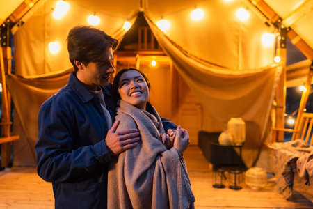 Man embracing asian girlfriend in blanket near blurred glamping house during weekend