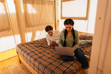 Asian freelancer using laptop near boyfriend with smartphone on bed in glamping houseの写真素材