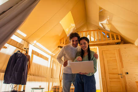 Man with cup hugging asian girlfriend pointing at laptop in glamping house