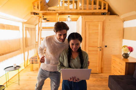Interracial couple with cup using laptop in glamping house during weekendの写真素材