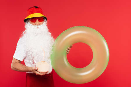 Santa claus in sunglasses holding swim ring and cocktail in coconut isolated on redの写真素材