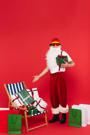 Santa claus in sunglasses pointing at presents on deck chair near shopping bags on red backgroundの写真素材