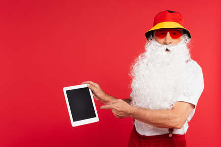 Santa claus in panama and sunglasses pointing at digital tablet isolated on redの写真素材