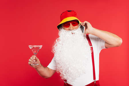 Santa claus in sunglasses talking on smartphone and holding cocktail isolated on redの写真素材