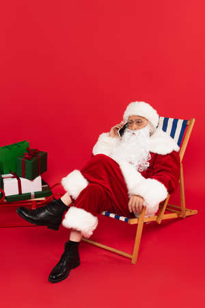 Santa claus talking on smartphone on deck chair near sleigh with gifts on red backgroundの写真素材