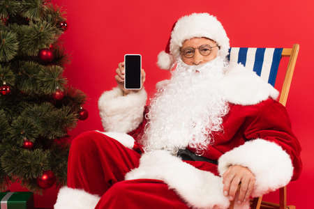 Santa claus holding smartphone on deck chair near christmas tree on red backgroundの写真素材
