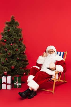 Santa claus holding cocktail in coconut on deck chair near christmas tree on red backgroundの写真素材