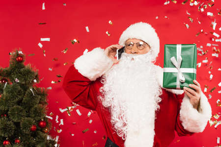 Santa claus talking on smartphone and holding gift under confetti near christmas tree on red...