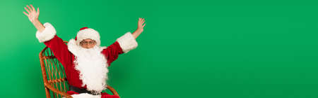 Excited santa claus sitting on wicker chair isolated on green, bannerの写真素材