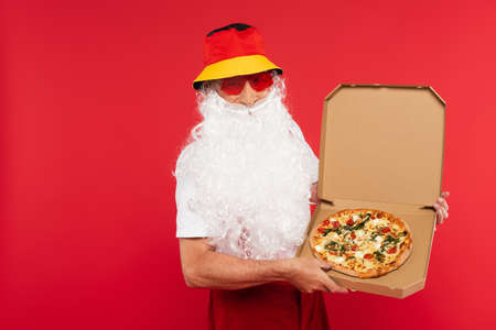 Santa claus in panama and sunglasses holding tasty pizza in box isolated on redの写真素材