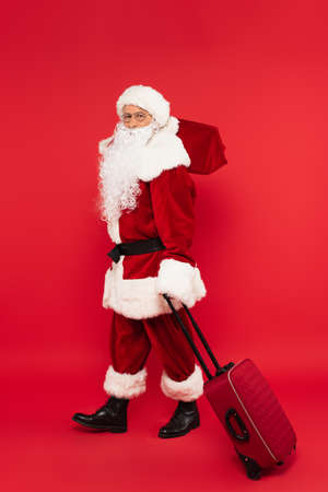 Santa claus in costume holding suitcase and sack on red backgroundの写真素材