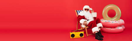 Santa in sunglasses sitting on deck chair near vintage boombox and swim rings on red background, bannerの写真素材