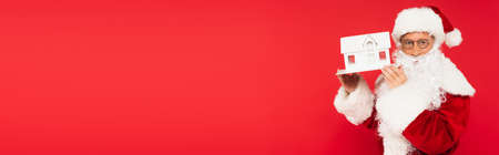 Santa claus in eyeglasses holding house model isolated on red, bannerの写真素材