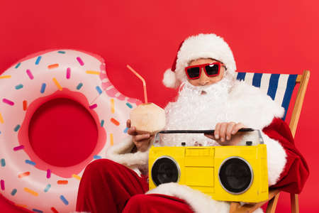 Santa in sunglasses holding cocktail in coconut and boombox on deck chair near swim ring on red backgroundの写真素材