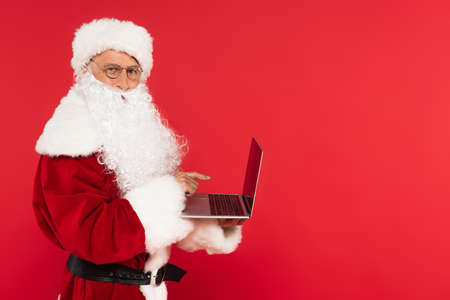 Santa claus in costume pointing at laptop with blank screen isolated on redの写真素材