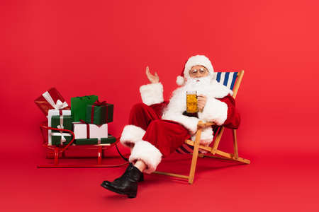 Santa claus holding glass of beer on deck chair near sleigh with presents on red backgroundの写真素材