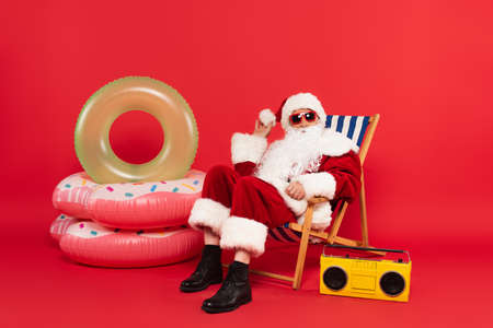 Santa claus in sunglasses and costume sitting on deck chair near swim rings and boombox on red backgroundの写真素材