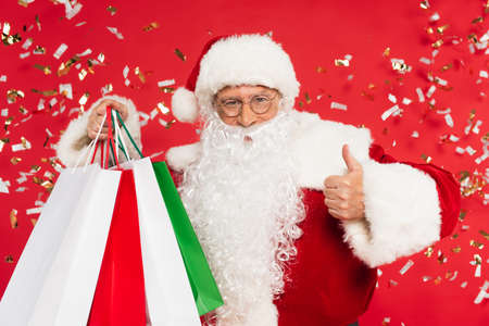 Santa claus showing like and holding shopping bags under confetti on red backgroundの写真素材
