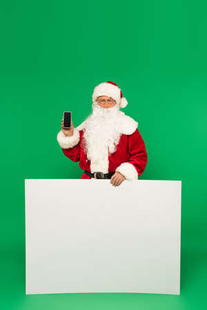 Santa claus holding smartphone with blank screen and placard on green backgroundの写真素材