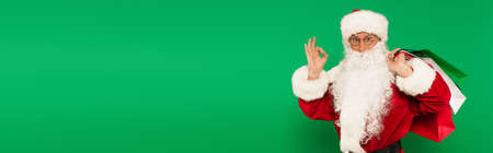 Bearded santa claus with shopping bags showing ok gesture isolated on green, bannerの写真素材
