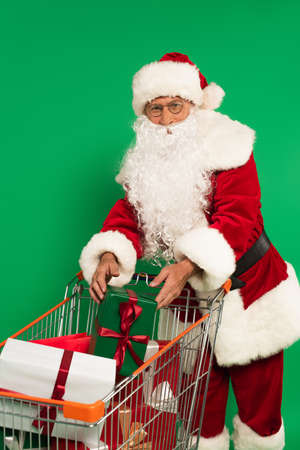 Santa claus in costume putting gift in shopping cart isolated on greenの写真素材