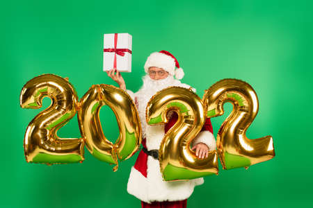 Santa claus holding gift near balloons in shape of 2022 numbers isolated on green