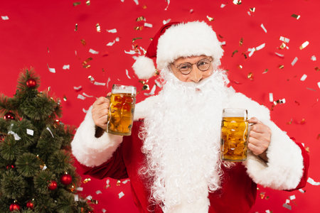 Santa claus holding glasses of beer near confetti and christmas tree on red backgroundの写真素材