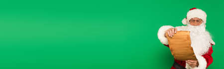 Santa claus in hat and eyeglasses holding paper isolated on green, banner