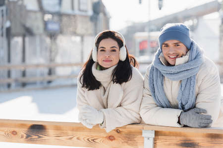 happy young woman in ear muffs and joyful man in winter hat leaning on wooden border on ice rinkの写真素材