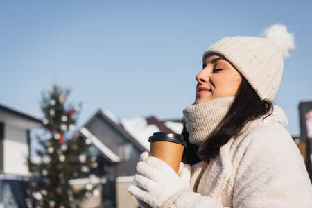 cheerful woman with closed eyes and winter hat holding paper cup outside