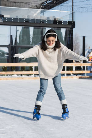 full length of young woman in sweater and ear muffs smiling while skating on ice rinkの写真素材