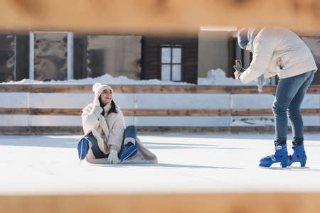 man in hat taking photo of girlfriend in ice skates in ice rink with blurred foregroundの写真素材