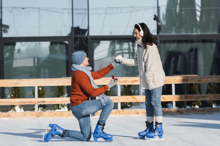 man holding box with wedding ring and kneeling while making proposal to happy woman in ear muffs on ice rinkの写真素材