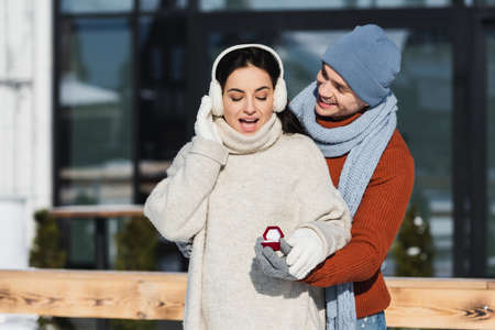 smiling man in winter hat holding box with wedding ring near amazed young woman in ear muffsの写真素材