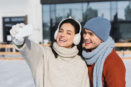 happy woman in sweater and ear muffs taking selfie with cheerful boyfriend on ice rinkの写真素材