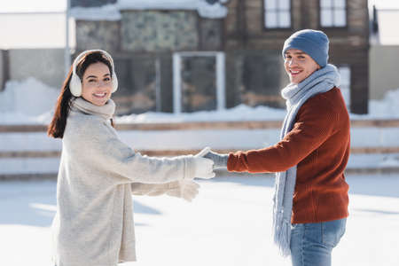 smiling young woman in ear muffs holding hands with cheerful boyfriend on ice rinkの写真素材