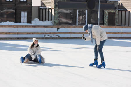 man in hat taking photo of happy girlfriend in ice skates on ice rinkの写真素材