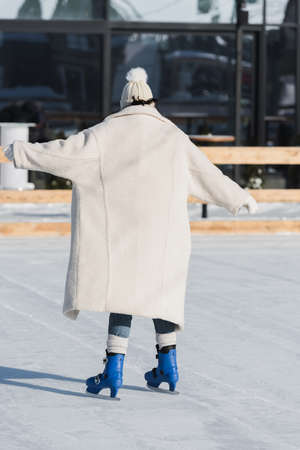 back view of young woman in winter hat and coat skating on ice rinkの写真素材