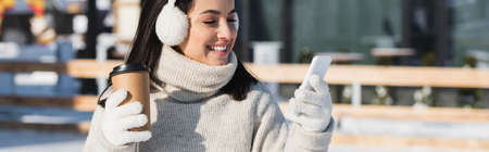 happy young woman in sweater and ear muffs holding paper cup and using smartphone on ice rink, bannerの写真素材