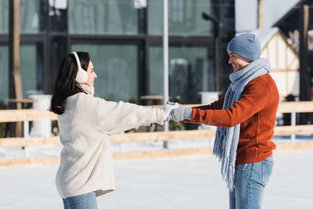 side view of positive young woman in ear muffs holding hands with boyfriend on ice rinkの写真素材