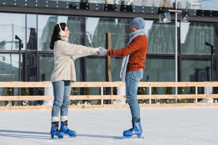 full length of young woman in ear muffs holding hands with boyfriend on ice rinkの写真素材