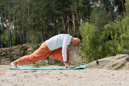 side view of buddhist in harem pants practicing crescent lunge pose on yoga mat in forestの写真素材
